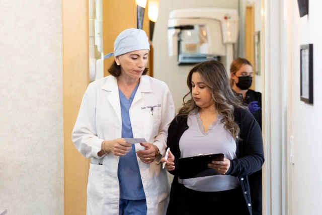 Dentist in white coat and cap discusses patient records with woman in clinic hallway.