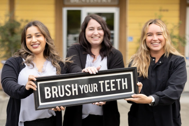 Three dental professionals holding "Brush Your Teeth" sign outside St. Helena Dental Studio