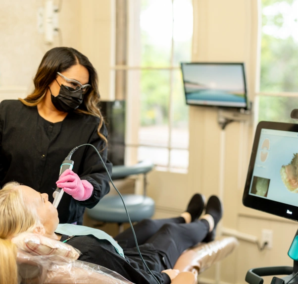 Dental hygienist in mask treats patient in modern dental office with monitors visible.