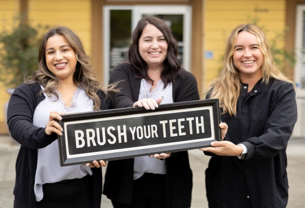 Three dental professionals holding "Brush Your Teeth" sign outside St. Helena Dental Studio building.